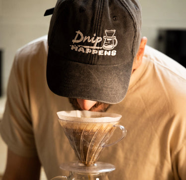 Person wearing a cap with 'Drip Happens' text, holding a coffee filter over a cup.