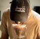 Person wearing a cap with 'Drip Happens' text, holding a coffee filter over a cup.