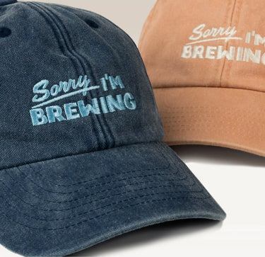 Two Sorry I'm Brewing Hats from RAVE COFFEE, made of soft brushed cotton—one in dark blue with blue embroidery, the other in light brown with white embroidery—each featuring the playful phrase “Sorry, I’m BREWING” embroidered on the front. These vintage-style caps offer subtly contrasting details for a unique statement.