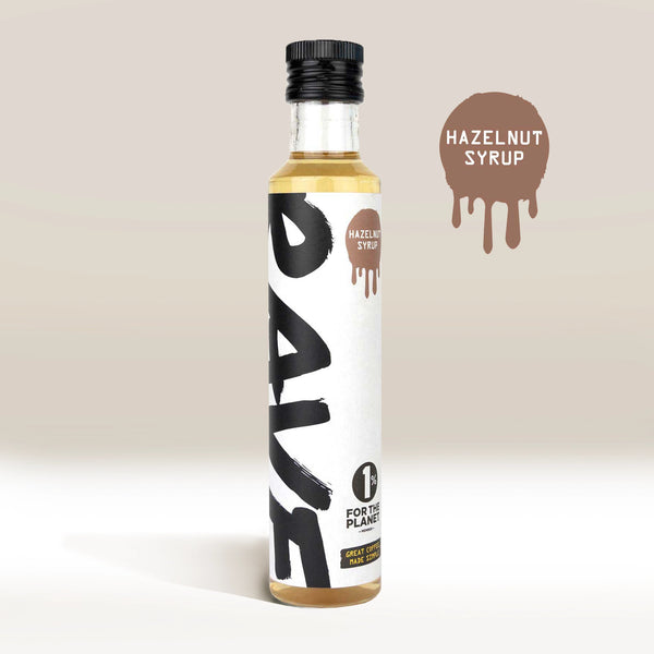 A clear bottle labeled RAVE COFFEE contains light brown Rave Coffee Syrups. A round "Hazelnut Syrup" graphic with dripping edges appears on the UK-blended coffee syrup, plus a 1% for the Planet badge at the label’s bottom.