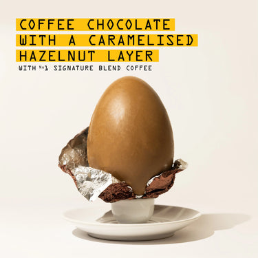 A large RAVE COFFEE Coffee Chocolate Easter Egg, caramel-colored, rests in a half-open foil wrapper on a white saucer. Above it, bold yellow and black text reads: 