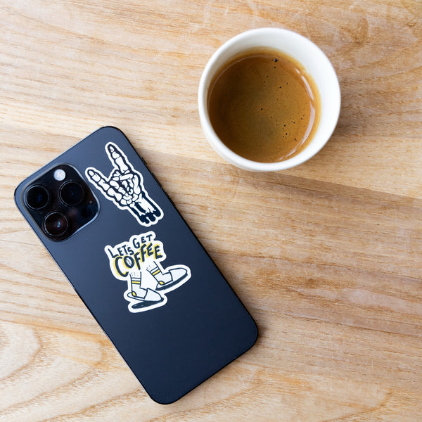 A cup of espresso sits on a wooden table next to a black smartphone decorated with stickers from the Sticker Pack Illustrated by Abigail Hudspith, featuring designs like a skeleton hand and one that says LETS GET COFFEE.