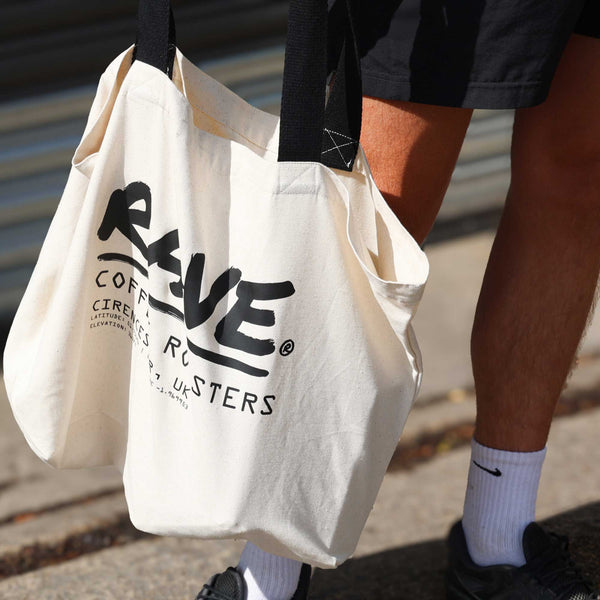 A person in black shorts and white socks carries the Rave Co-ordinates Tote Bag—an eco-friendly recycled canvas tote with black straps and "REVE Coffee Roasters" text—set against a metal shutter and pavement.