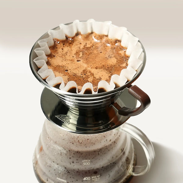 A Kalita Wave 185 Steel Coffee Maker by Kalita, with ground coffee in a paper filter, sits over a glass carafe as freshly brewed coffee drips below and the bloom forms a foamy layer on top.
