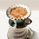 A Kalita Wave 185 Steel Coffee Maker by Kalita, with ground coffee in a paper filter, sits over a glass carafe as freshly brewed coffee drips below and the bloom forms a foamy layer on top.