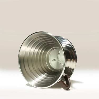 The Kalita Wave 185 Steel Coffee Maker by Kalita lies on its side, showing its ribbed interior and round base, with a brown handle to the right against a light beige background.