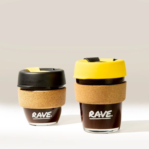 Two Rave Coffee Glass KeepCups by KeepCup, one with a black lid and one yellow, both featuring cork sleeves and filled with coffee, sit side by side on a light surface with "RAVE." printed on each.