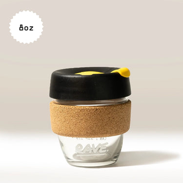 A KeepCup Rave Coffee Glass KeepCup, featuring durable tempered glass, a cork band, black lid, and yellow stopper, sits against a light beige background with 