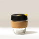 The KeepCup Rave Coffee Glass KeepCup features tempered glass, a cork band, black plastic lid with yellow tab, and “RAVE” printed on the glass. Set against a plain light background.