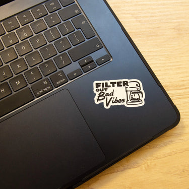 A black laptop with a RAVE COFFEE Drip Happens Sticker Pack featuring a FILTER OUT Bad Vibes vinyl and a minimalist coffee maker line art. Ideal for fans of unique coffee stickers. The laptop is on a light wood surface.