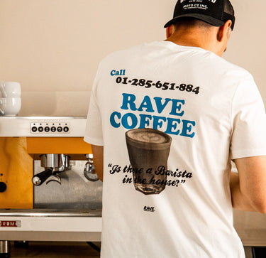 Barista wearing a white graphic t-shirt with 'RAVE COFFEE' printed in blue and black, featuring a coffee cup illustration and the phrase 'Is there a Barista in the house?'. He is standing in front of a coffee machine.