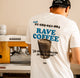 Barista wearing a white graphic t-shirt with 'RAVE COFFEE' printed in blue and black, featuring a coffee cup illustration and the phrase 'Is there a Barista in the house?'. He is standing in front of a coffee machine.