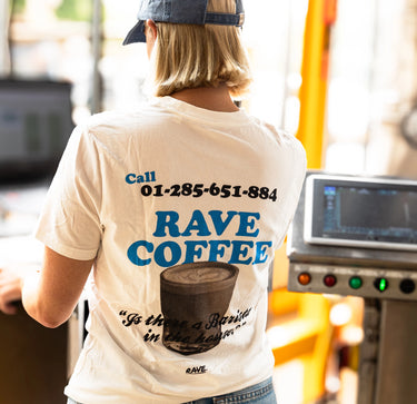 A person viewed from behind wearing a cream-colored t-shirt with a large blue and black graphic on the back that reads 'Call 01-285-651-884 RAVE COFFEE' and features an illustration of a coffee cup. The person is also wearing a blue baseball cap in a brightly lit, slightly blurred indoor setting.