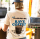 A person viewed from behind wearing a cream-colored t-shirt with a large blue and black graphic on the back that reads 'Call 01-285-651-884 RAVE COFFEE' and features an illustration of a coffee cup. The person is also wearing a blue baseball cap in a brightly lit, slightly blurred indoor setting.
