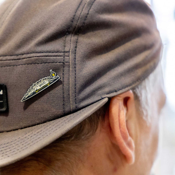 A close-up side view of a person wearing a gray cap with the Rave Alligator Pin Badge attached above the ear, a distinctive RAVE Coffee accessory.