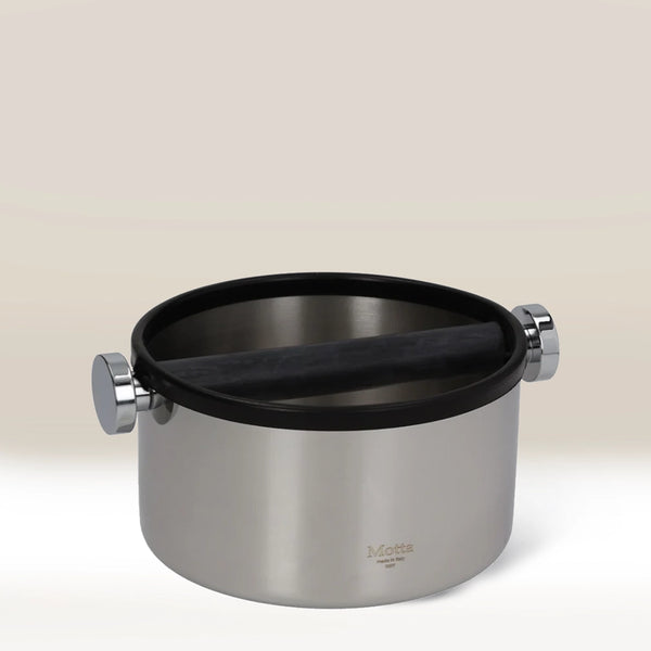 The MOTTA Knock Box 165mm features a sturdy stainless steel build with a black rubber bar for easy grounds disposal. The MOTTA logo appears on the front, set against a gradient beige background.