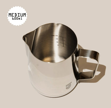 The Crema Pro Milk Steaming Jug or Pitcher holds 20 oz (600ml/0.6 Litre), features a handle and internal measurements, and is ideal for latte art. The stainless steel jug is shown on a light beige background, labeled MEDIUM 600ml.