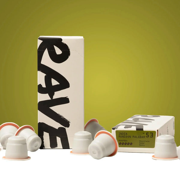 Indian Monsoon Malabar Coffee Pods are scattered around two RAVE boxes on a greenish-yellow gradient. One box stands upright; the other lies flat showing details of these Nespresso compatible coffee pods.