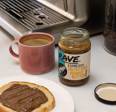 A jar of Rave Espresso Salted Date Peanut Butter sits on a white countertop next to a pink coffee mug, a slice of toast spread with peanut butter, and a coffee machine in the background.