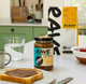 A jar of RAVE Coffee Espresso Salted Date Peanut Butter—an indulgent, vegan-friendly, and palm oil-free treat—rests on a kitchen counter beside toast spread with espresso peanut butter, a glass of water, a mug, a bowl of raspberries, and a bag of coffee blend.
