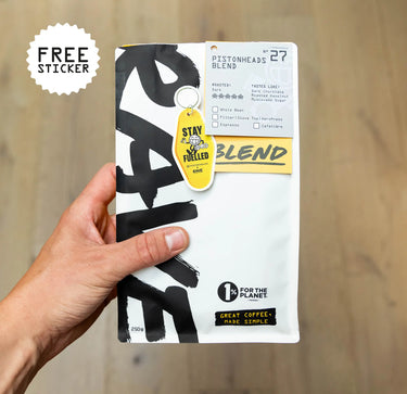 A hand holds a bag of RAVE COFFEE's Nº 27 Pistonheads Blend limited-edition coffee, featuring bold black lettering, a yellow Stay Fueled keychain, and a label offering a free sticker. Text highlights that 1% of sales support the planet.