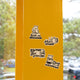 The Drip Happens Sticker Pack by RAVE COFFEE features four vinyl stickers—Sorry, I’m brewing bear, Hot stuff mug, Filter out bad vibes coffee maker, and Drip happens dripper—on a yellow pole with trees and cars in the background.