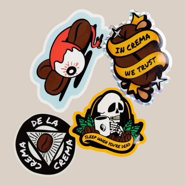 The Rave Tattoo Sticker Pack by RAVE COFFEE features four holographic coffee-themed stickers: a coffee bean with an eyeball, “IN CREMA WE TRUST” ribbon, skull with “SLEEP WHEN YOU’RE DEAD,” and “CREMA DE LA CREMA” triangle.