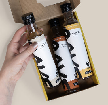 A hand holds one of three small glass bottles from a RAVE Coffee Pick N’ Mix Syrups Set, packaged in a cardboard box and labeled Vanilla, Hazelnut, and Caramel.