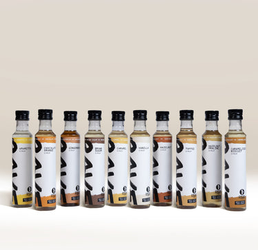 Ten glass bottles of RAVE Coffee Pick N’ Mix Syrups Set in a row, each with a white label featuring the flavor name and a large black brushstroke graphic.