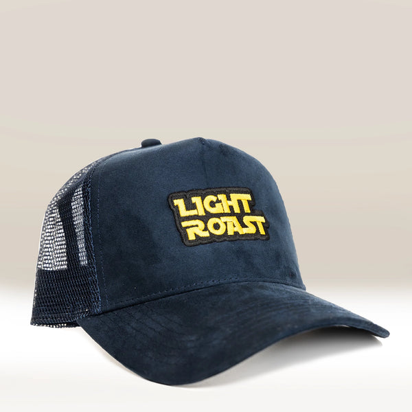 A dark blue Suede Trucker Cap from RAVE COFFEE featuring a yellow and white embroidered front patch that reads LIGHT ROAST in bold block letters, set against a smooth gradient background.