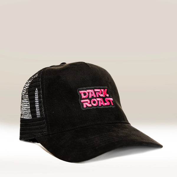 The Suede Trucker Cap by RAVE COFFEE is a black retro-style hat with a mesh back and "DARK ROAST" embroidered in bold pink and white on the front, shown against a light beige background.