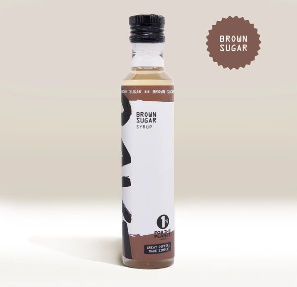 A bottle of Rave Coffee Syrups by RAVE COFFEE, featuring a black cap and labeled with BROWN SUGAR SYRUP and 1% for the Planet, sits against a beige background. A starburst graphic in the top right corner reads BROWN SUGAR, highlighting this UK-made flavoured coffee syrup.