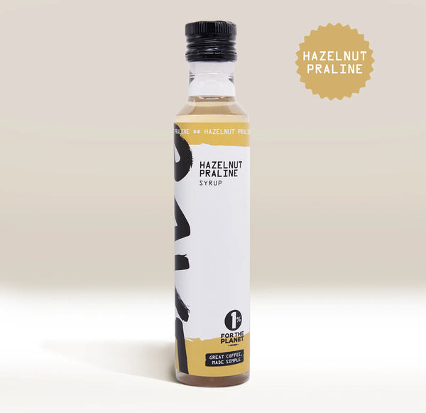 A bottle of Rave Coffee Syrups by RAVE COFFEE, featuring a black cap, white and gold label, and the 1% for the Planet emblem. The “Hazelnut Praline” text is highlighted in a tan circle. This UK-made flavoured coffee syrup is perfect for adding rich, nutty sweetness to your favourite coffee syrups collection.