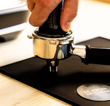 A close-up of a hand using a black espresso tamper to press coffee grounds into a stainless steel portafilter resting on a black mat.