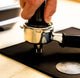 A close-up of a hand using a black espresso tamper to press coffee grounds into a stainless steel portafilter resting on a black mat.