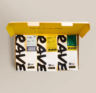 Three Ravecoffee bags from the Best Selling Coffee Bundle Gift Pack, each filled with premium medium/dark roast beans, are neatly arranged in an open yellow gift box. Each bag displays a bold black 