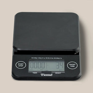 The Tiamo Timer Scale is a black coffee scale with a flat platform, built-in timer, “Start/Stop” and “On/Off/Tare” buttons, and a display for time and weight. The Tiamo brand name appears below the screen.