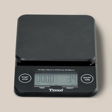 The Tiamo Timer Scale is a black coffee scale with a flat platform, built-in timer, “Start/Stop” and “On/Off/Tare” buttons, and a display for time and weight. The Tiamo brand name appears below the screen.