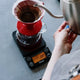 A person pours hot water into a red pour-over dripper with a paper filter, set on a glass carafe and the Tiamo Timer Scale by Tiamo, which displays both brewing time and weight.