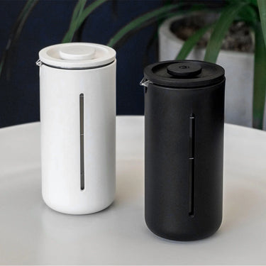 Two sleek, cylindrical soap dispensers—one white, one black—sit side by side on a light surface, echoing the design of the Timemore French Press U by Timemore. Each has a slim vertical window to show contents, with a green plant blurred in the background.