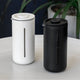 Two sleek, cylindrical soap dispensers—one white, one black—sit side by side on a light surface, echoing the design of the Timemore French Press U by Timemore. Each has a slim vertical window to show contents, with a green plant blurred in the background.