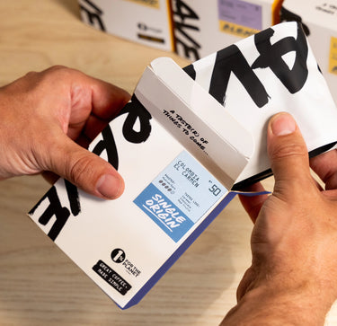 A person’s hands opening a white coffee box labeled “Best Selling Single Origins - Whole Bean Variety Pack” from Ravecoffee with a blue label and bold black text, revealing whole bean coffee on a light wooden surface with more Ravecoffee boxes in the background.