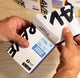 A person’s hands opening a white coffee box labeled “Best Selling Single Origins - Whole Bean Variety Pack” from Ravecoffee with a blue label and bold black text, revealing whole bean coffee on a light wooden surface with more Ravecoffee boxes in the background.