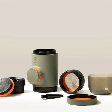 A WACACO Minipresso GR2 travel coffee maker with detachable cup, filters, lids, and brush is neatly arranged on a light surface against a neutral background.