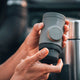 A person holds a WACACO Minipresso GR2, a gray portable espresso maker travel cup with a textured lid and circular logo. The background is slightly blurred, revealing part of a metal container.