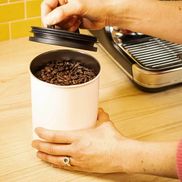 A person opens a Planetary Design White Airscape Coffee Storage Container filled with coffee beans on a wooden countertop, with a coffee machine visible in the background.