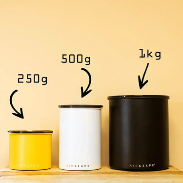 Three Planetary Design Airscape Coffee Storage Containers—yellow, white, and black—labeled 250g, 500g, and 1kg sit on wood. Each uses Airscape technology to remove oxygen and keep your coffee fresher for longer.