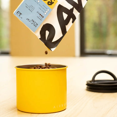 Whole coffee beans are poured from a tilted bag into a Planetary Design Yellow Airscape Coffee Storage Container on a light wooden table, with a coiled black cord and large windows in the background.