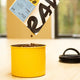 Whole coffee beans are poured from a tilted bag into a Planetary Design Yellow Airscape Coffee Storage Container on a light wooden table, with a coiled black cord and large windows in the background.