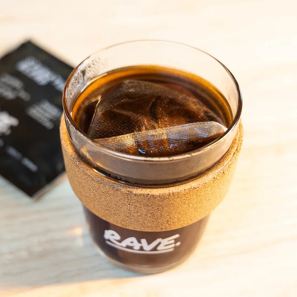 A glass of dark coffee with a cork band labeled RAVE sits on a light wooden surface, containing a steeping Colombia El Carmen Compostable Coffee Bag from RAVE COFFEE. In the background is a black packet featuring the specialty coffee single serve sachets 10 pack.